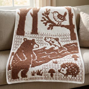 May include: A white crochet blanket featuring a brown woodland pattern. The design includes a bear standing near a river with a jumping fish, a bird perched on a tree branch, a hedgehog, and small mushrooms, all rendered in a textured stitch.