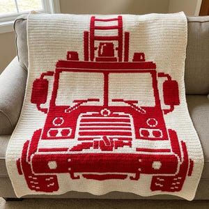 May include: A rectangular crochet blanket featuring a large, centered red and cream graphic of a fire truck. The design displays the front view of the vehicle, including a ladder on top, windshields, headlights, and a front bumper.