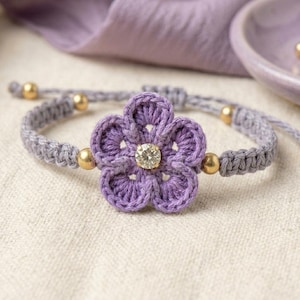 May include: A lavender crochet flower bracelet featuring a central clear crystal stone. The band is crafted from grey knotted cord and accented with four small gold-toned metal beads positioned symmetrically around the floral centerpiece.