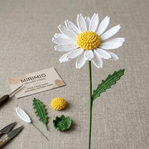以下が含まれることがあります： 白い花びらと黄色の中心、緑の茎と葉を持つ手作りのかぎ針編みのデイジー。追加のかぎ針編みの作品には、黄色の中心、葉、小さな緑色のベースが含まれます。画像には、「MIRIMIO CROCHET STUDIO WILD FIELD DAISY（REVISED EDITION 2026）」と書かれた名刺も表示されています。