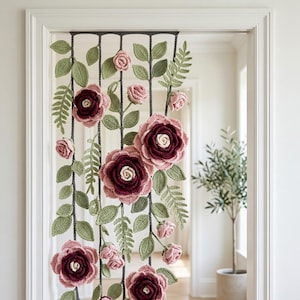 Puede incluir: Una cortina floral de ganchillo hecha a mano. La cortina presenta grandes rosas burdeos y rosas, capullos rosas más pequeños y hojas verdes. Las flores y las hojas están unidas a cuerdas grises verticales, creando una cubierta decorativa para puertas o ventanas.