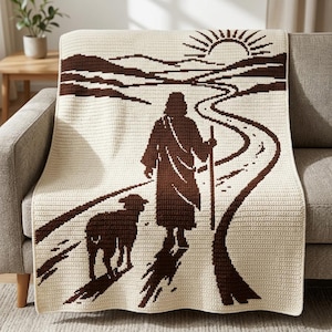 May include: A cream-colored crochet blanket featuring a dark brown graphic design of a figure in a robe holding a staff, walking along a winding path with a sheep nearby. The background depicts rolling hills and a rising sun with radiating rays.