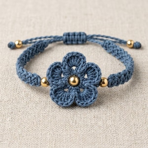 Puede incluir: Una pulsera de ganchillo azul con un diseño de flor y un centro dorado. La pulsera tiene una banda trenzada con cuentas doradas. El cierre de macramé ajustable permite un ajuste cómodo.