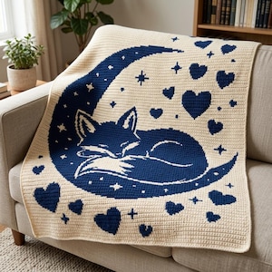 Op de afbeelding: Een crèmekleurige gehaakte deken met een grote donkerblauwe maansikkel waarin een slapende vos is opgerold. Het ontwerp is versierd met verspreide donkerblauwe hartjes en kleine vierpuntige sterren over het gehele oppervlak van het textiel.