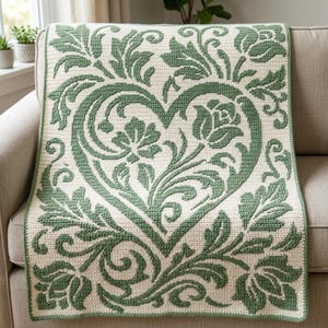 May include: A rectangular crochet blanket featuring a cream-colored background with a prominent sage green heart motif at the center. The design includes intricate floral and leafy scrollwork patterns that extend from the heart to the edges.