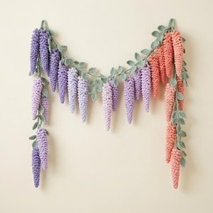Pode incluir: Uma guirlanda decorativa com cachos de flores de crochê semelhantes a glicínias em tons de roxo, lavanda e coral, conectados por uma trepadeira de crochê verde contínua com pequenas folhas. Os elementos florais pendem em cascata.