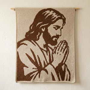 Puede incluir: Un tapiz de pared de ganchillo rectangular con un diseño gráfico bicolor en marrón y crema de una persona con pelo largo y barba, representada de perfil con las manos juntas en posición de oración, suspendido de una varilla de madera.