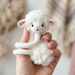 May include: A handmade white crochet lamb toy with a curly wool-like texture. The lamb has pink inner ears, a small black eye, and a smiling face. The neck is long and coiled, and the toy is held in a hand.