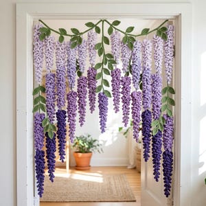 May include: A crocheted wisteria garland hangs in a doorway. The garland features cascading strands of purple flowers in varying shades, from light lavender to deep violet. Green leaves and vines are interspersed throughout the design, creating a naturalistic look.