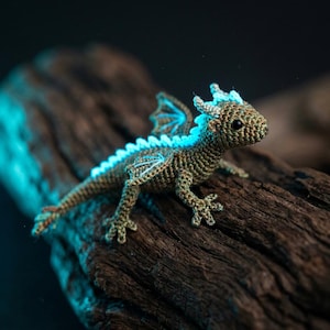 May include: A crocheted dragon figurine in shades of brown and green, with blue accents on its wings and spine. The handmade dragon is posed on a piece of dark wood, showcasing its detailed craftsmanship. The dragon's size is approximately 6 inches long.