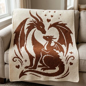 May include: A cream-colored crochet blanket featuring a large brown dragon and a smaller dragon sitting together. The design includes decorative hearts and star shapes surrounding the two dragons, created with a contrasting brown yarn pattern.