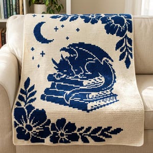 Può includere: Una coperta all'uncinetto color crema con un disegno grafico blu scuro che raffigura un drago che dorme su una pila di libri. Il motivo include una luna crescente, piccole stelle e grandi decorazioni floreali negli angoli, in filato blu.