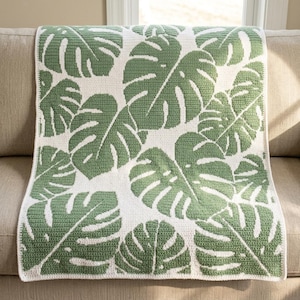 May include: A rectangular crochet blanket featuring a repeating pattern of large, green monstera leaves against a solid white background. The textile has a visible stitched texture throughout, with a clean, finished border along all four edges.