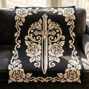 May include: A rectangular crochet blanket featuring a black background with a contrasting cream-coloured design. The pattern displays a central sword oriented vertically, flanked by detailed rose motifs, all enclosed within a floral border.