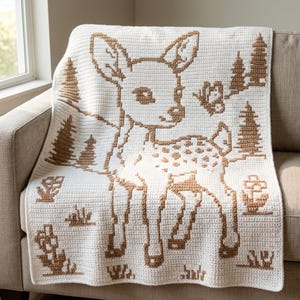 May include: A white crochet blanket featuring a brown deer motif in the center. The design includes brown pine trees, a small butterfly, and flowers surrounding the deer. The pattern is created with a textured stitch, displaying a woodland scene.
