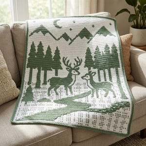 Puede incluir: Una manta de ganchillo rectangular con fondo blanco y un patrón en verde salvia. El diseño muestra una escena forestal con dos ciervos, pinos y picos de montañas bajo una luna creciente y estrellas, con un borde verde a juego.