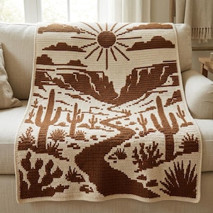 May include: A cream-coloured crochet blanket featuring a brown desert landscape design. The pattern displays a central sun, rocky canyon formations, a winding path, and various cacti including saguaro and prickly pear plants across the surface.