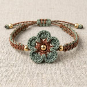 May include: A bracelet featuring a central crocheted flower in sage green and brown yarn with a gold-toned bead at its center. The band is made of braided sage green and brown cord, accented with small gold-toned beads and an adjustable sliding knot.