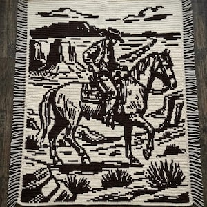 May include: A rectangular crochet blanket featuring a two-tone dark brown and cream design. The pattern depicts a rider on horseback in a desert landscape with mesas and cacti. The blanket is finished with fringe along the left and right sides.