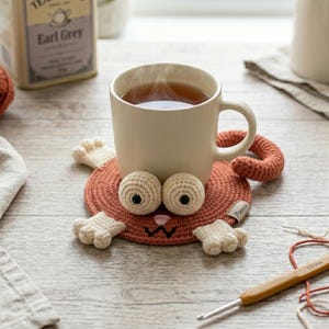 May include: A beige mug filled with tea sits on a handmade, orange cat-shaped coaster. The coaster features crocheted eyes, paws, and a tail. A crochet hook and tea box are in the background.