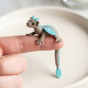 Puede incluir: Una pequeña figura de dragón de ganchillo en tonos beige y azul. El dragón tiene alas detalladas y una larga cola, y está posado en un dedo. Esta criatura de fantasía hecha a mano es un artículo decorativo único.