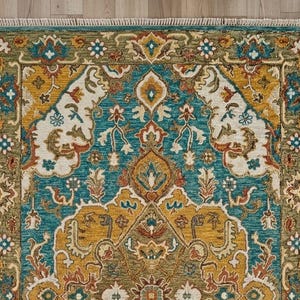 May include: A detailed close-up of a rectangular rug with an intricate pattern. The rug features a teal, gold, and cream color palette with floral and geometric designs. The border is a darker green with a fringed edge.