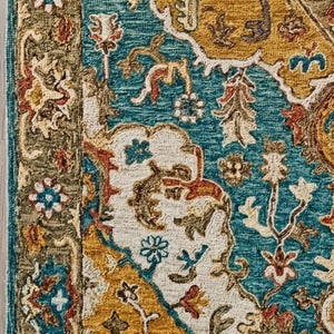 May include: A close-up of a patterned rug with a teal, gold, and cream color scheme. The rug features an intricate floral design with a border of olive green and teal. The rug has a fringe along the bottom edge.