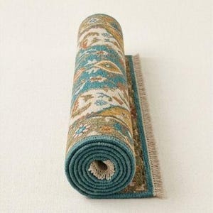 May include: Rolled-up area rug with a teal border and a detailed pattern in shades of teal, beige, orange, and yellow. The rug has a fringed edge and is displayed against a white background. The rolled portion reveals the rug's intricate design.