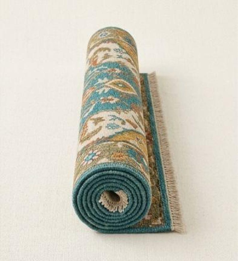 May include: Rolled-up area rug with a teal border and a detailed pattern in shades of teal, beige, orange, and yellow. The rug has a fringed edge and is displayed against a white background. The rolled portion reveals the rug's intricate design.