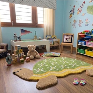 May include: A plush, turtle-shaped area rug in shades of green, yellow, and tan, featuring a white floral pattern on its shell. The rug is positioned on a wooden floor in a room containing a white play table, a toy storage unit, and a teddy bear.