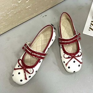 May include: A pair of white Mary Jane flats with black polka dots. The shoes have red trim, straps with buckles, and a bow detail. The interior features a quilted pattern.