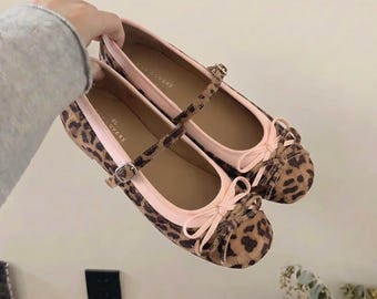 May include: A pair of leopard print ballet flats with a light pink trim and bow detail. The shoes have a strap with a buckle closure. The shoes are held up by a hand.