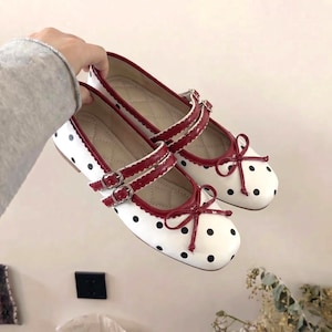 Peut inclure: Ballerines Mary Jane blanches à pois noirs, bordées de rouge et ornées de nœuds. Les chaussures ont des brides doubles avec des boucles argentées. Les semelles sont beige clair.