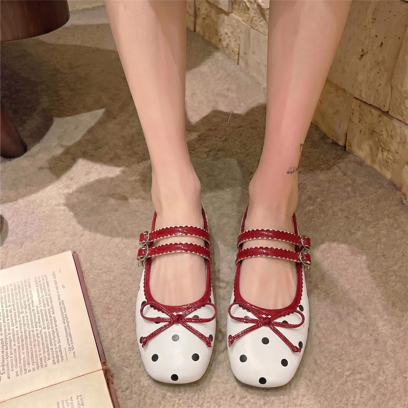 May include: White ballet flats with black polka dots and red trim. The flats have a double strap design with buckles and a bow detail on the toe. The shoes are designed for comfort and style.