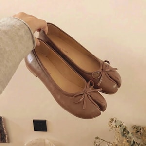 Op de afbeelding: Een paar bruine leren ballerina's met een split-teenontwerp en kleine strikaccenten. De schoenen hebben een ronde neus en een platte zool. De merknaam "MDA Shoes" is zichtbaar op de binnenzool.