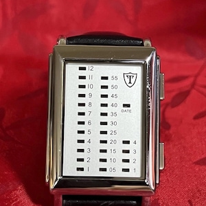 May include: A rectangular silver wristwatch with a black leather band. The watch face is white with black indicators for hours and minutes. The watch has a shield logo with the letter "T" and the word "DATE".