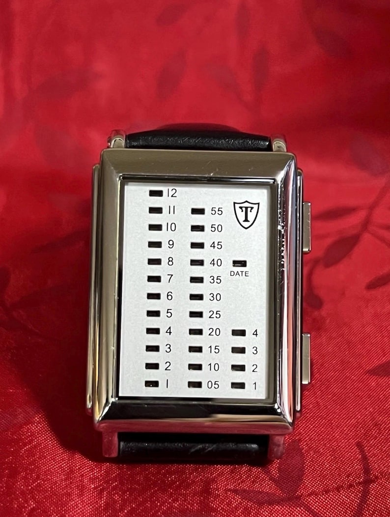 May include: A rectangular silver wristwatch with a black leather band. The watch face is white with black indicators for hours and minutes. The watch has a shield logo with the letter "T" and the word "DATE".