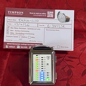 May include: A silver Detomaso watch with a black leather band. The watch face displays a digital time with green and blue lights. A Timpson service card is in the background, with details about the watch.