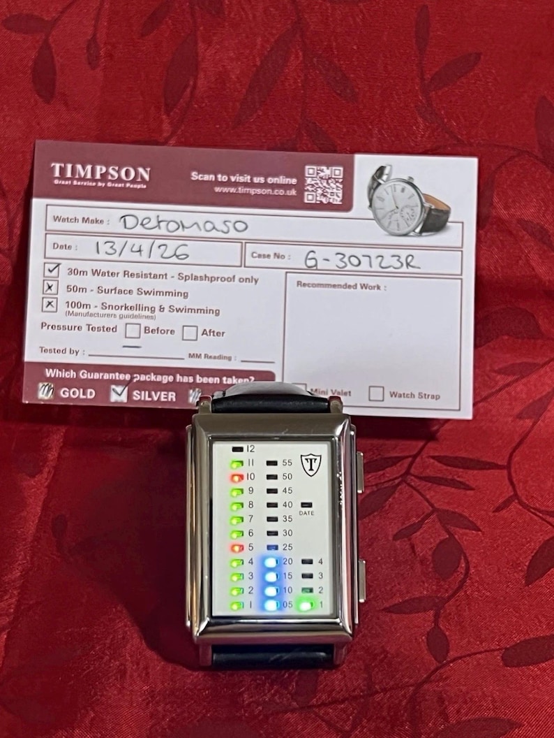 May include: A silver Detomaso watch with a black leather band. The watch face displays a digital time with green and blue lights. A Timpson service card is in the background, with details about the watch.