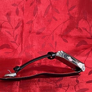 May include: A black leather bracelet with a silver clasp and a rectangular metal accent piece. The metal piece is engraved with the word "DETOMASO". The bracelet is displayed on a red textured background.