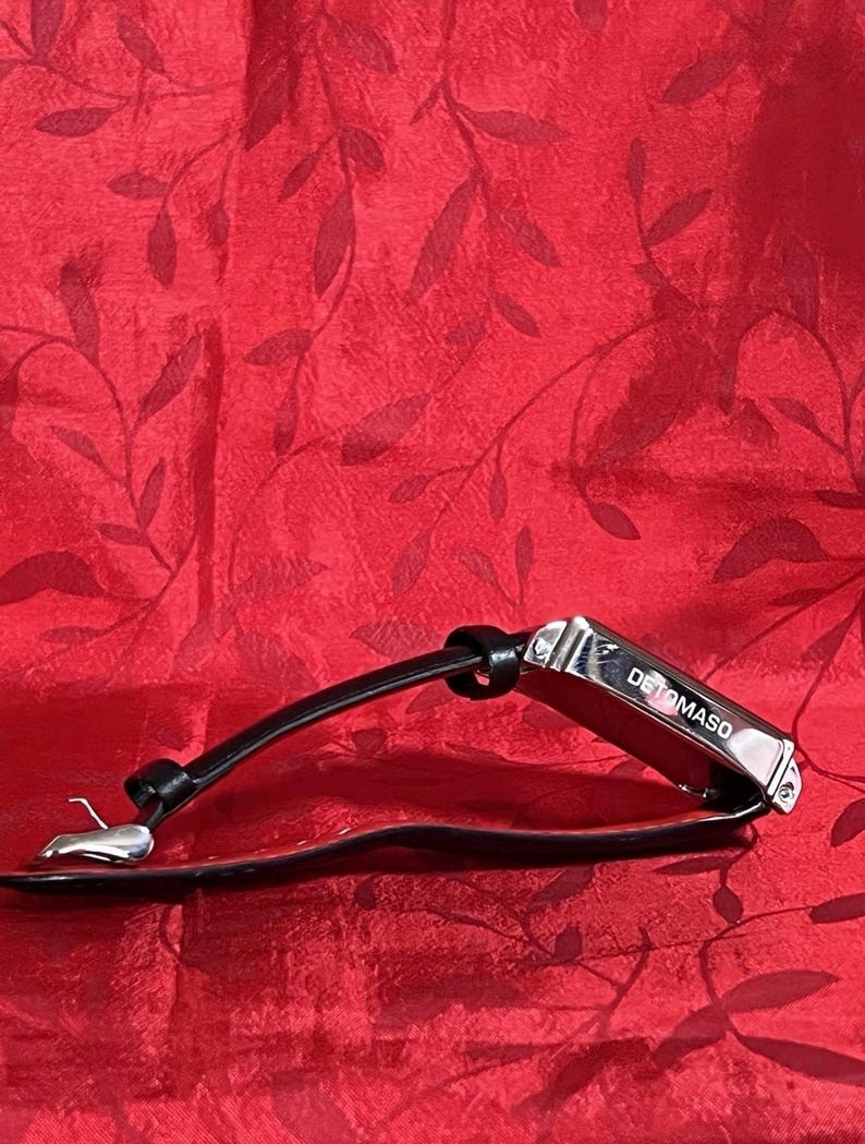 May include: A black leather bracelet with a silver clasp and a rectangular metal accent piece. The metal piece is engraved with the word "DETOMASO". The bracelet is displayed on a red textured background.