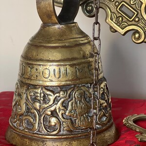 May include: Antique brass bell with intricate relief carvings and the inscription "D: QUI ME". A chain is attached to the bell, which is mounted on a decorative brass bracket. The bell sits on a red surface.