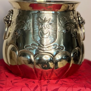 May include: A polished brass planter with a wide rim and a rounded base. The planter has decorative handles and a crest with a star and crescent moon. It sits on a red surface.