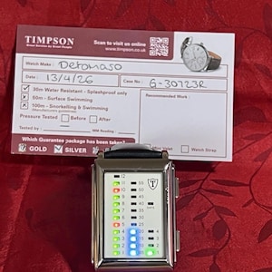 May include: A silver Detomaso watch with a black leather band, featuring a digital display with green and blue lights. The watch face displays numbers and a shield logo. A Timpson service card is in the background.