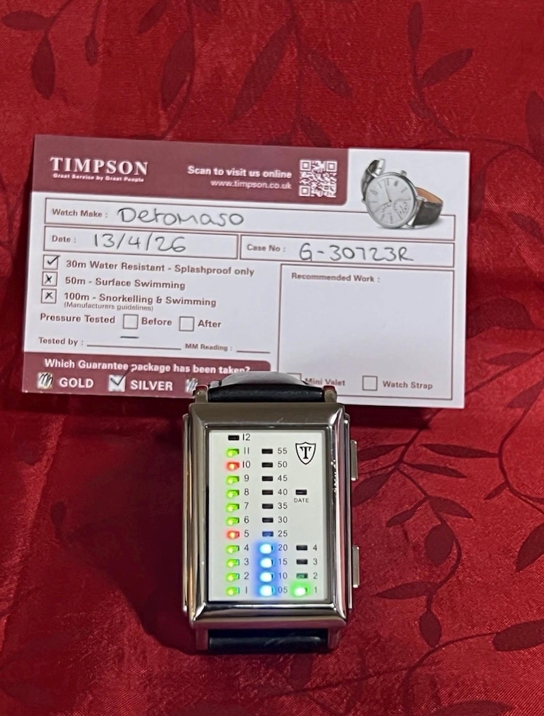 May include: A silver Detomaso watch with a black leather band, featuring a digital display with green and blue lights. The watch face displays numbers and a shield logo. A Timpson service card is in the background.