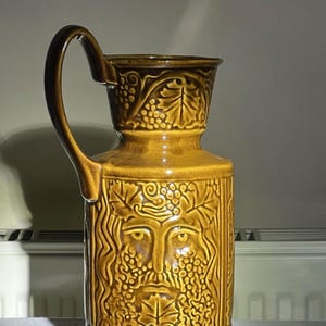 Peut inclure: Un pichet en céramique émaillée brune présentant un motif en relief d'un visage entouré de feuilles de vigne et de grappes. Le récipient possède un corps cylindrique haut, un col évasé et une anse incurvée sur le côté.