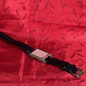 May include: A rectangular digital watch with a silver-colored case and a black leather band. The watch face is light pink with small, evenly spaced dots. The band has a buckle closure. The watch is displayed on a red background.