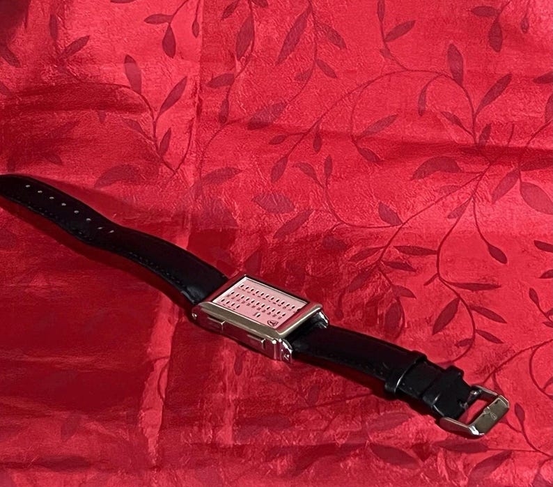 May include: A rectangular digital watch with a silver-colored case and a black leather band. The watch face is light pink with small, evenly spaced dots. The band has a buckle closure. The watch is displayed on a red background.
