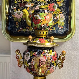 Könnte beinhalten: Ein schwarzer Metall-Samowar mit einer passenden Teekanne darauf, beide verziert mit leuchtenden, handgemalten Blumenmustern in Rot, Rosa, Weiß und Grün. Der Samowar verfügt über verzierte goldfarbene Griffe, einen zentralen Hahn und einen Sockel.