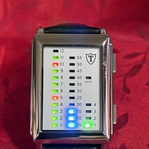 May include: A rectangular digital watch with a silver-colored case and a black leather band. The watch face displays time with a series of colored lights and numbers. The watch has the letter "T" inside a shield logo.